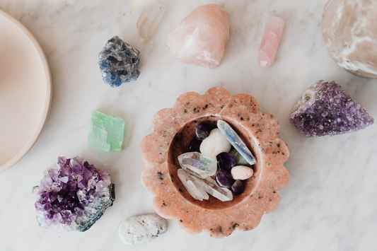 A flat lay of various healing crystals and stones arranged in a circle around a terracotta bowl on a white marble surface, with soft ambient lighting.