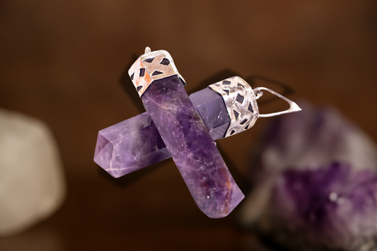 Two faceted amethyst crystal pendants with intricate silver caps, crossed elegantly against a warm blurred background with crystals in soft focus.